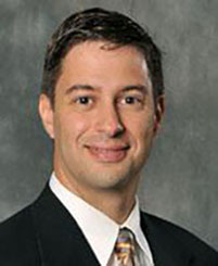 Spiro Maroulis headshot - fair-skinned man, smiling, brown hair, white collared shirt, black blazer, orange tie
