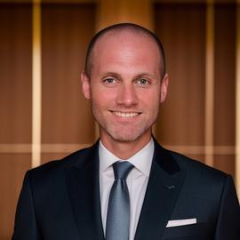 Anthony Howell headshot - fair-skinned man, smiling, brown hair, navy blazer, white collared shirt, blue tie