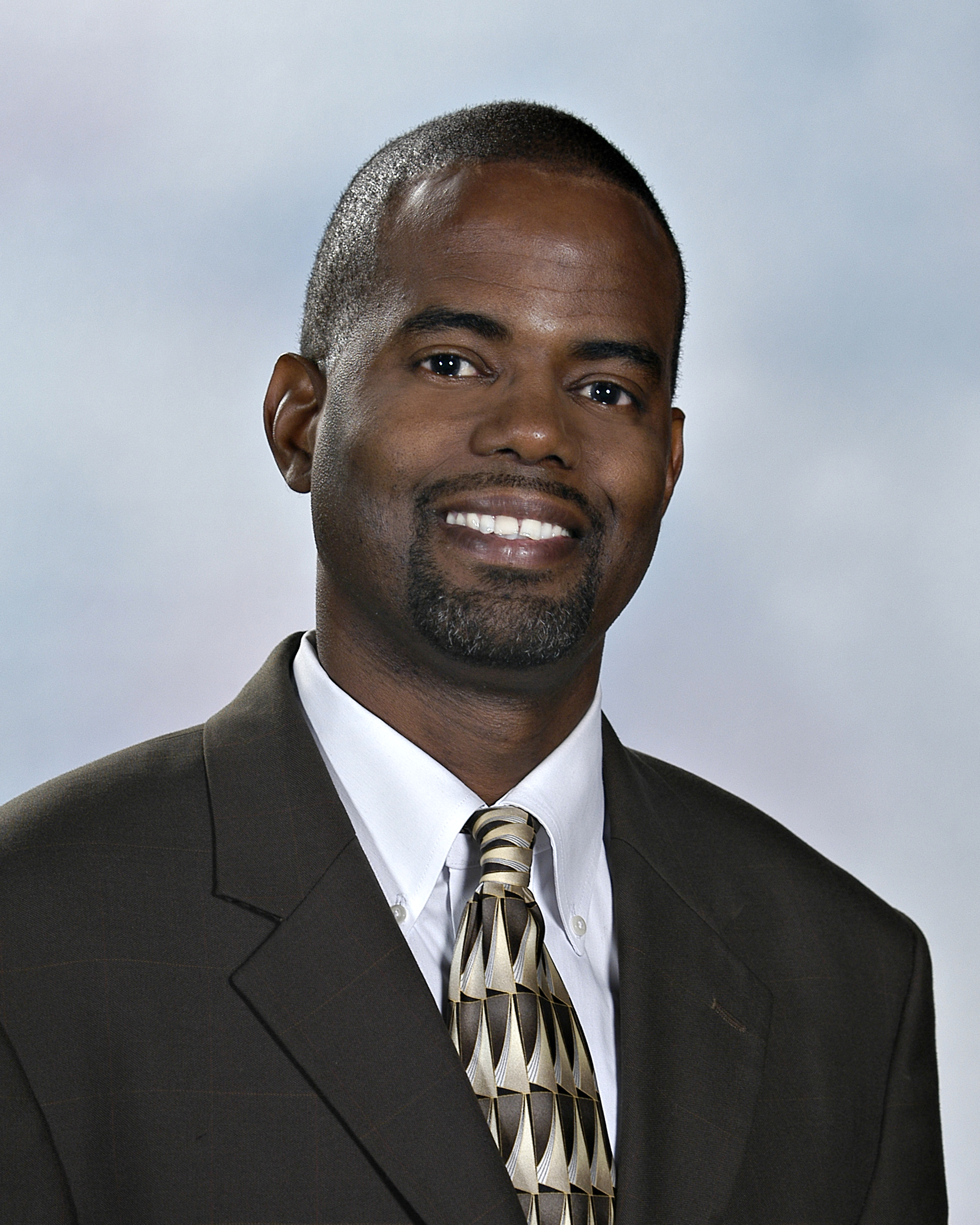 Jerry Hart headshot - dark-skinned man, smiling, black hair, white collared shirt, brown suit, brown patterned tie