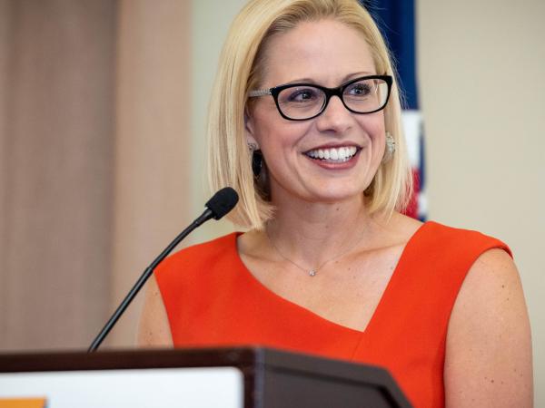 Senator Kyrsten Sinema. White Woman. Late 40s. Red dress. Black glasses.