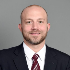Justin Stritch headshot - fair-skinned man, smiling, brown hair and beard, white collared shirt, navy blazer, red tie