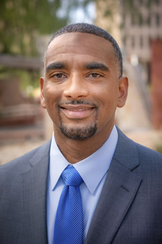 Jeff Barton headshot - dark-skinned man, smiling, black and gray hair and beard, light blue collared shirt, gray blazer, blue tie