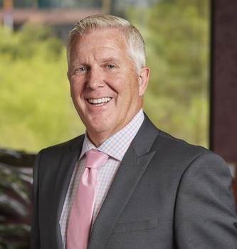 Kevin Phelps headshot - fair-skinned man, smiling, white hair, blue, pink, and white collared shirt, gray blazer, pink tie