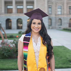﻿Claudia Sanson headshot - fair-skinned woman, smiling, black hair, maroon graduation cap, white dress, colorful and gold stoles, necklace